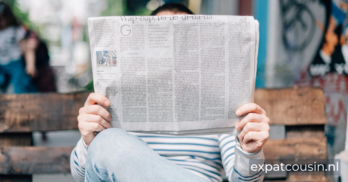 A man sitting on a wooden slatted bench reading the news paper. The paper is covering his face with only his head sticking out above. His hands a visible holding the paper sitting with his legs crossed wearing jeans and a white sweater with blue horizontal lines.