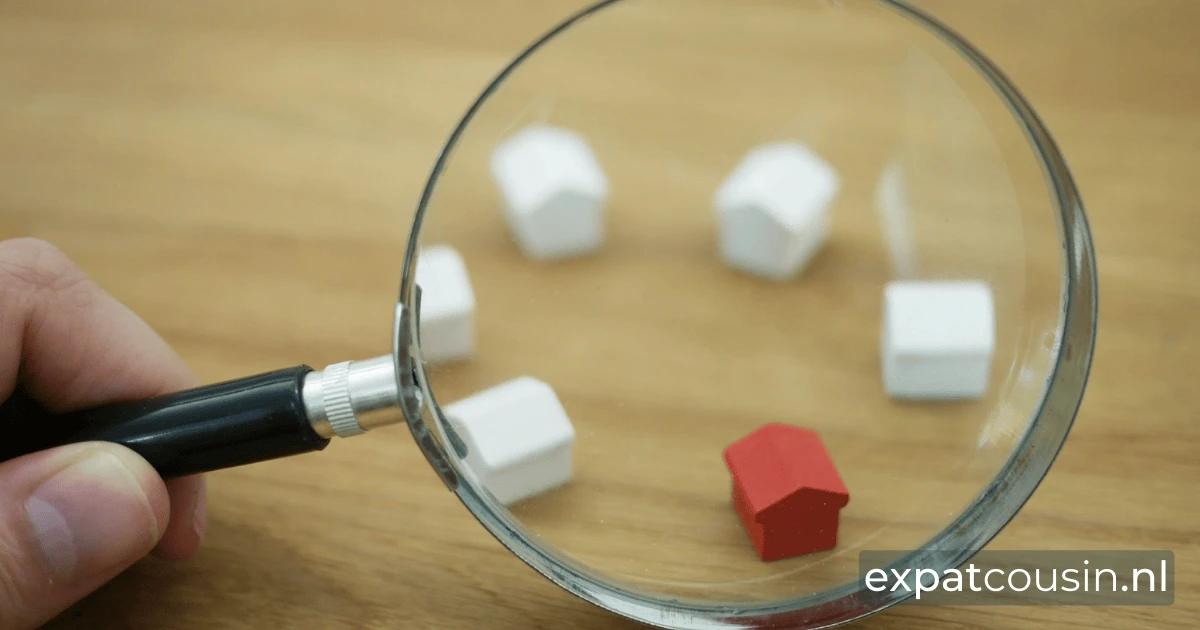A hand holding a magnifying glass which is magnifying a circle of 6 little toy houses, one red and the rest are white on a light wooden desk.