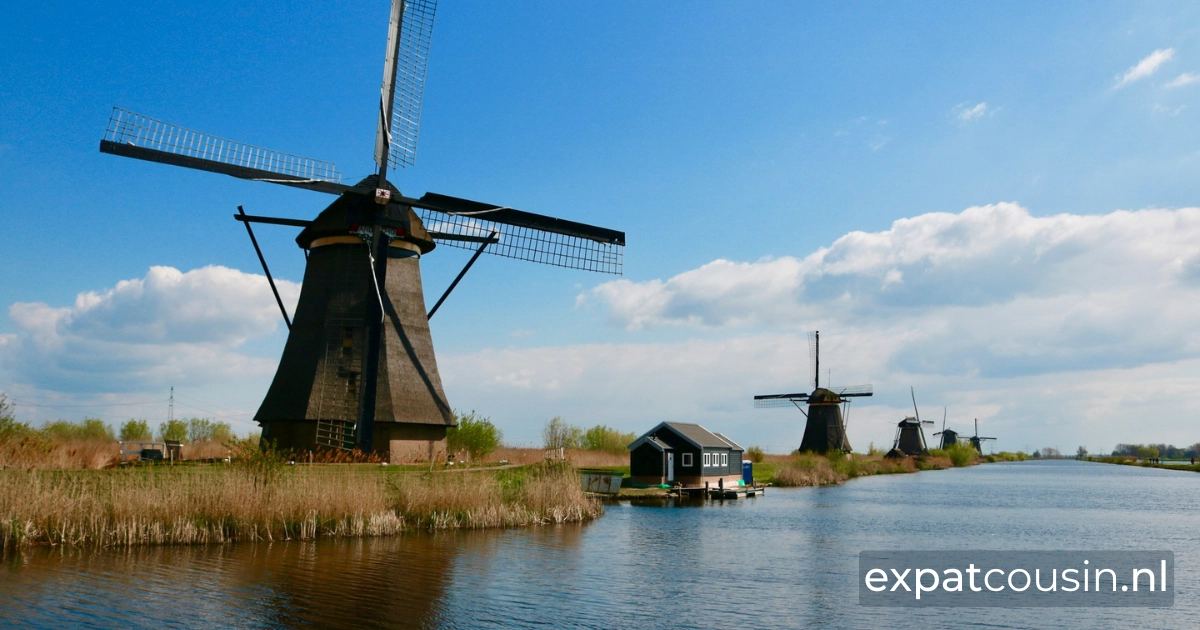 The kinderdijk in the netherlands landscape with a view of a large canal with five old windmills along the bank. As you look down the canal the windmills appear smaller and smaller in the distance. The water is calm and faint shadows of the windmill reflect on the water. The sky is blue with thin white clouds in the distance.