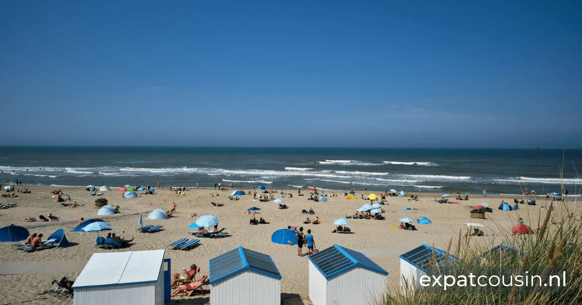 A beautiful summer day at the beach with blue roofed little white houses closeby. The large sandy beach is populated with people and umbrellas with the blue sea water in the distance.