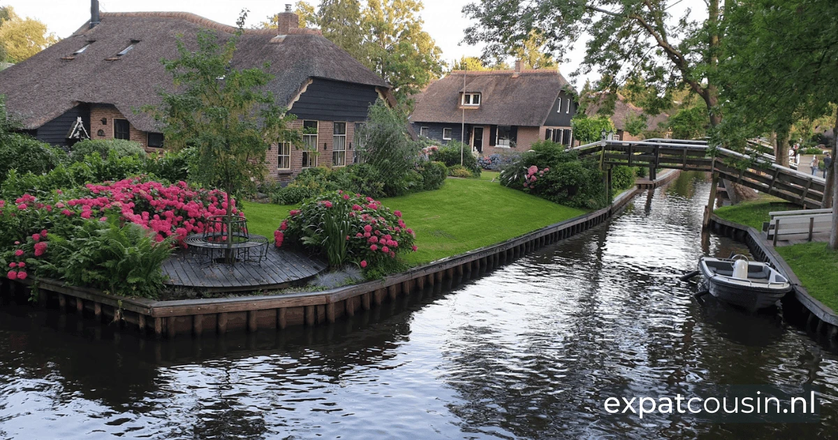 A netherlands canal and bridge with traditional thatched redbrick houses in the background. The houses have manicured green lawns with large shrubs and red flowers