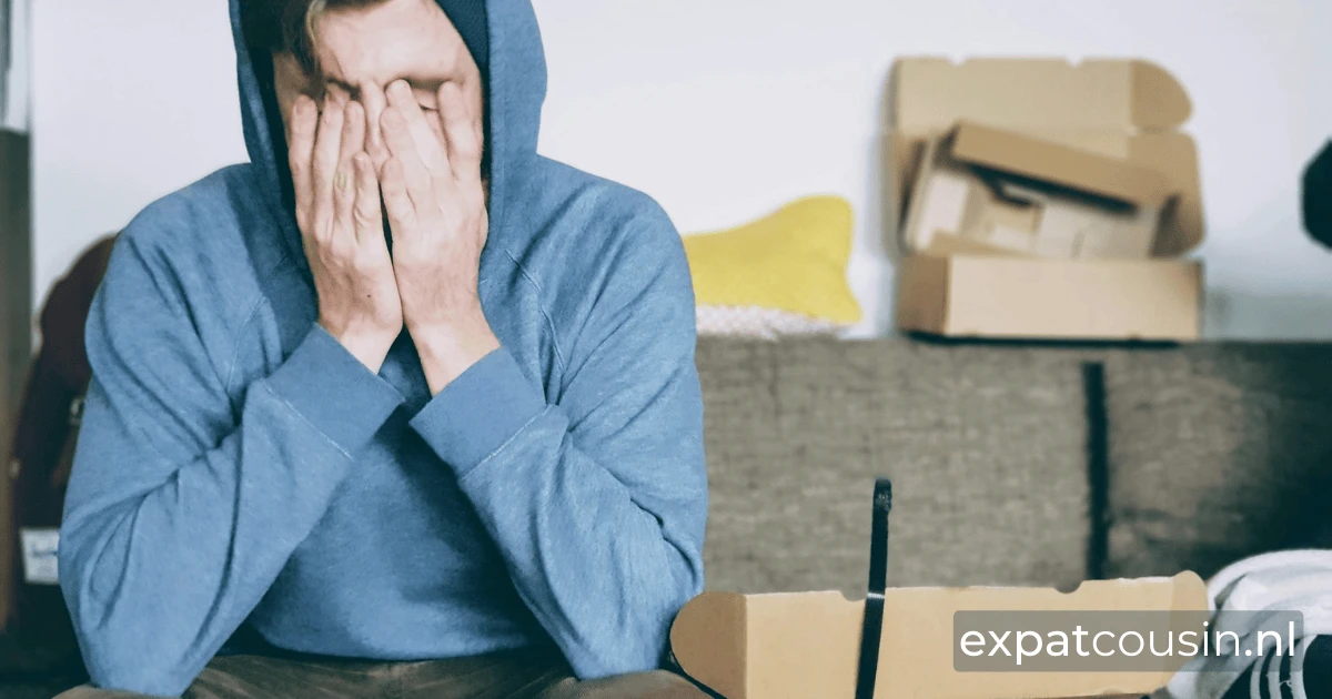 A young man with a blue hoody sitting with his hands over his face. An open box next the to the man and more unopened boxes in the background.